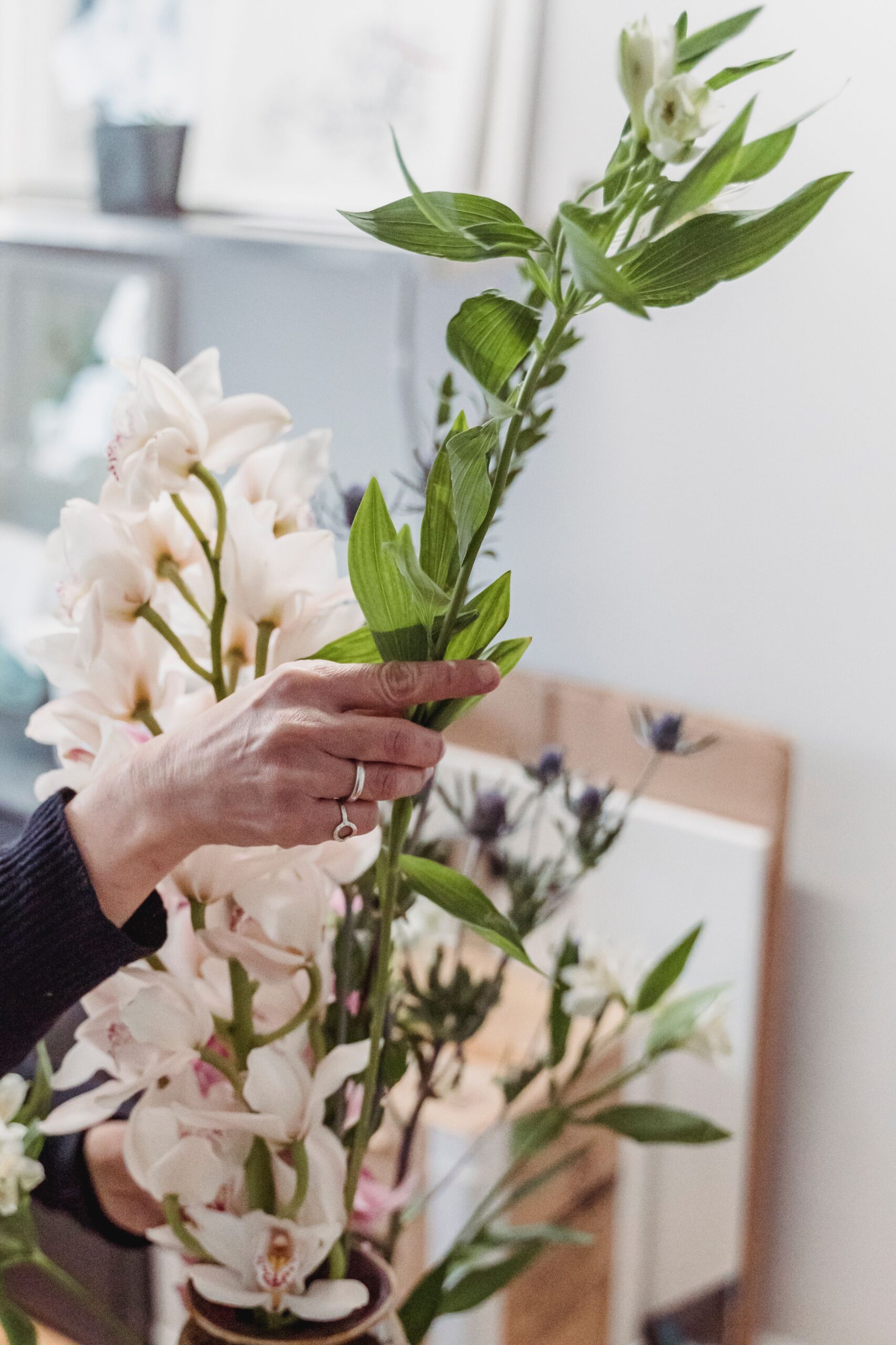 Hands arranging fresh flowers in a vase indoors, focusing on orchid blossoms and green leaves.